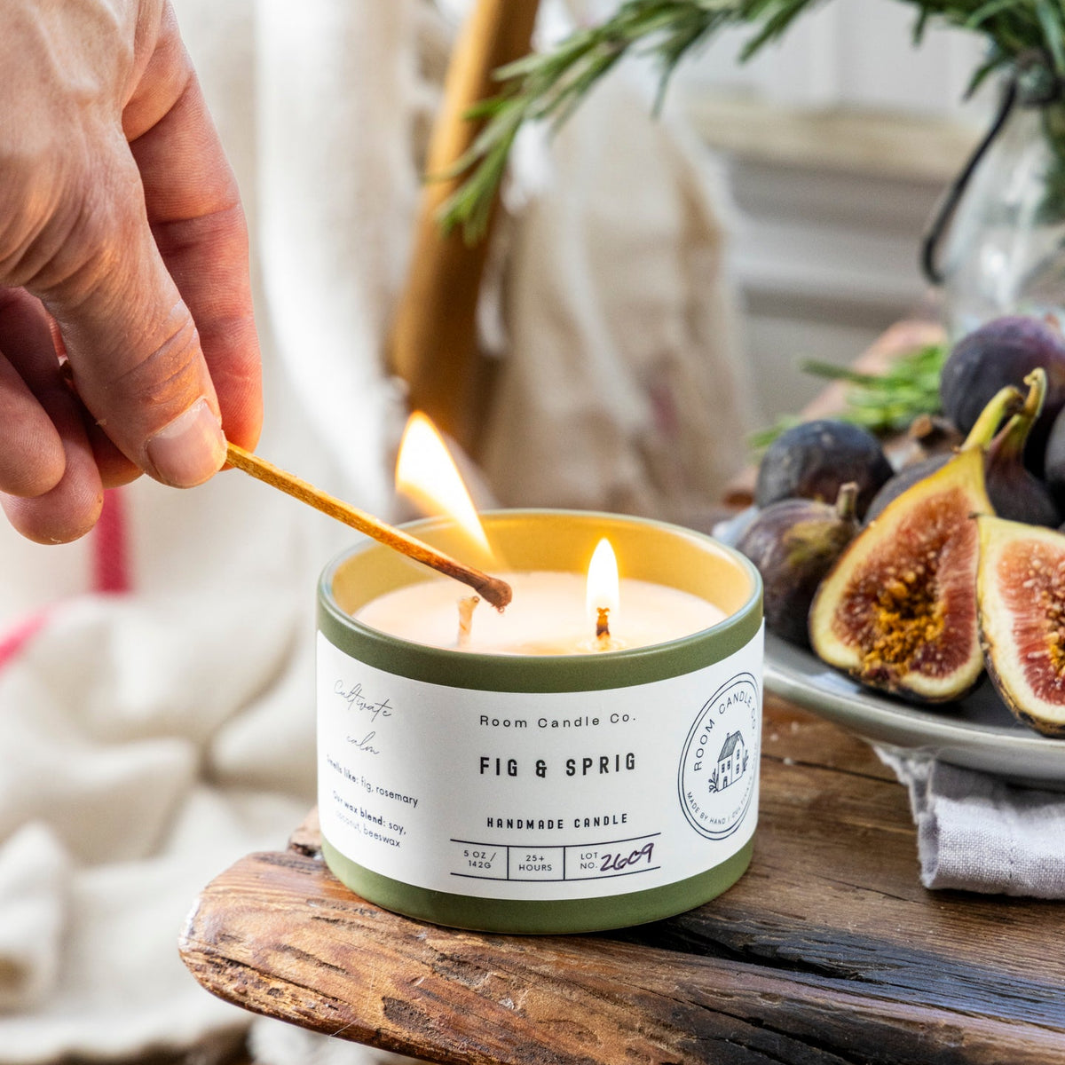 A hand lighting the Fig & Sprig candle by Room Candle Co. in a green ceramic vessel, styled with fresh figs and sprigs of rosemary.