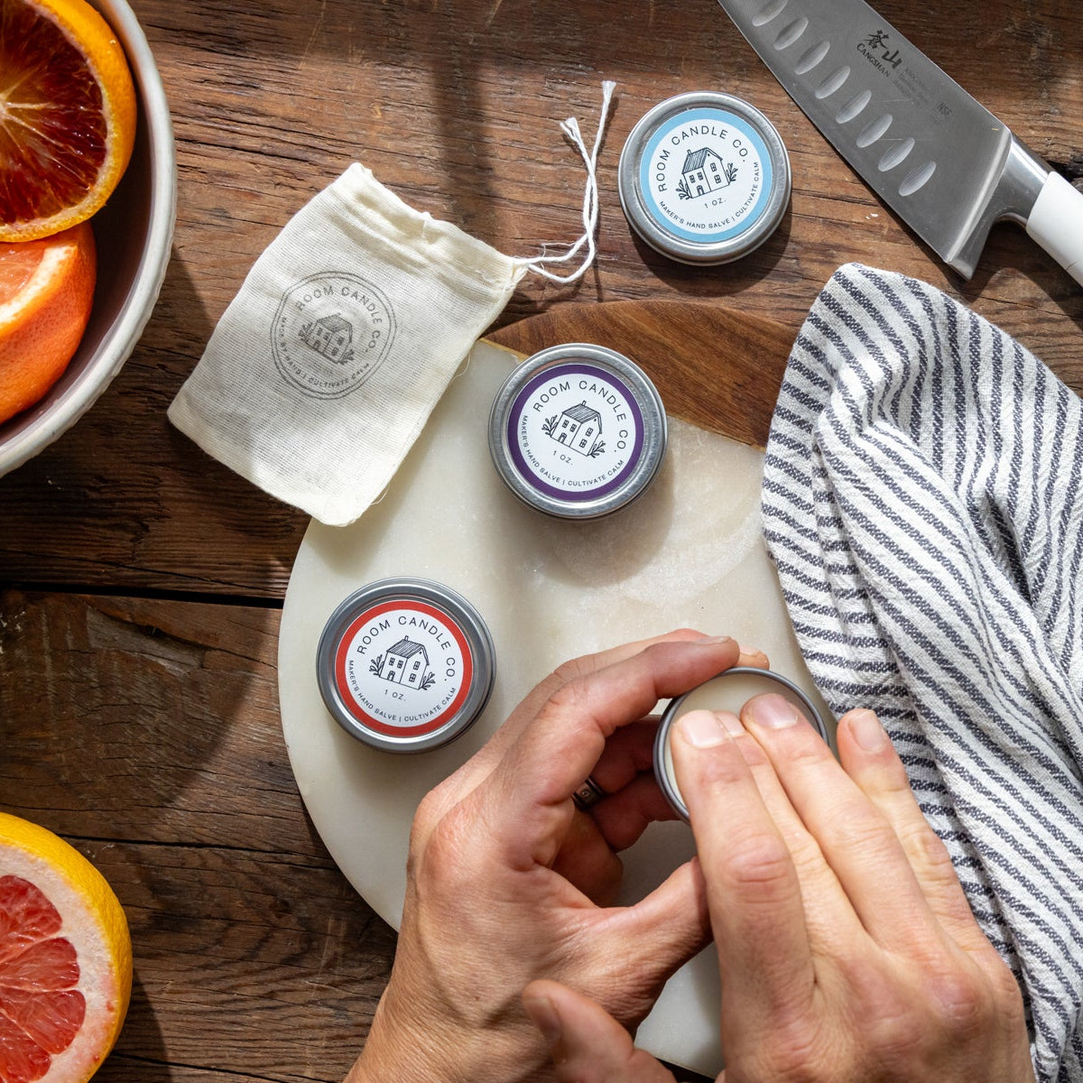A hand applying Room Candle Co. Maker’s Salve from a metal tin on a wooden cutting board alongside fresh citrus and a knife.