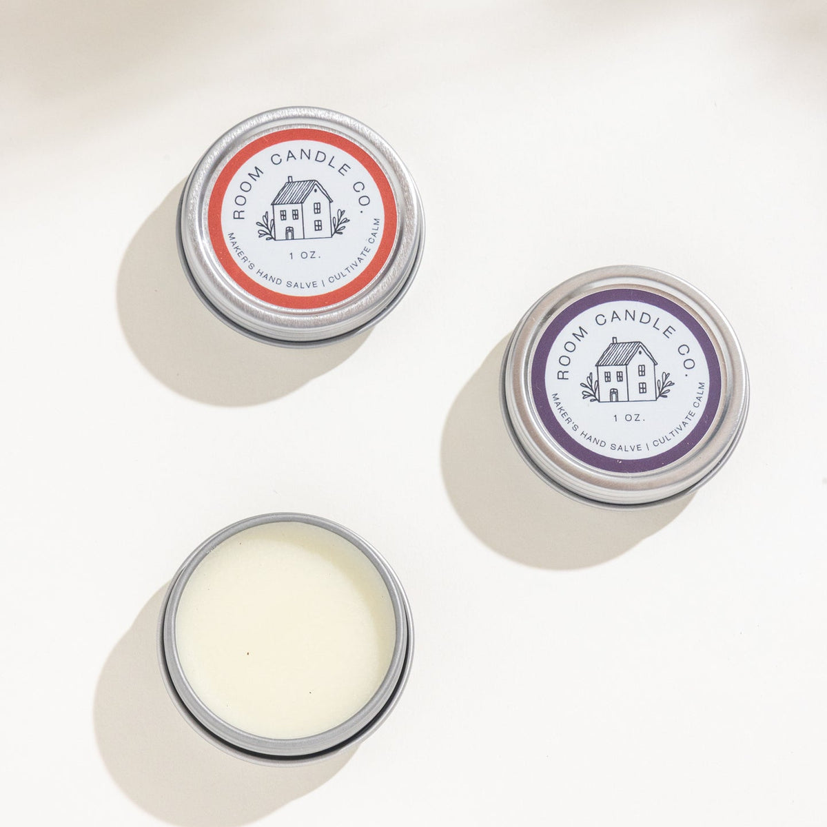 Three 1 oz metal tins of Room Candle Co. Maker's Hand Salve in Lemongrass and Tea Tree, Blood Orange and Myrrh, and Black Currant and Absinthe, displayed against a plain background.