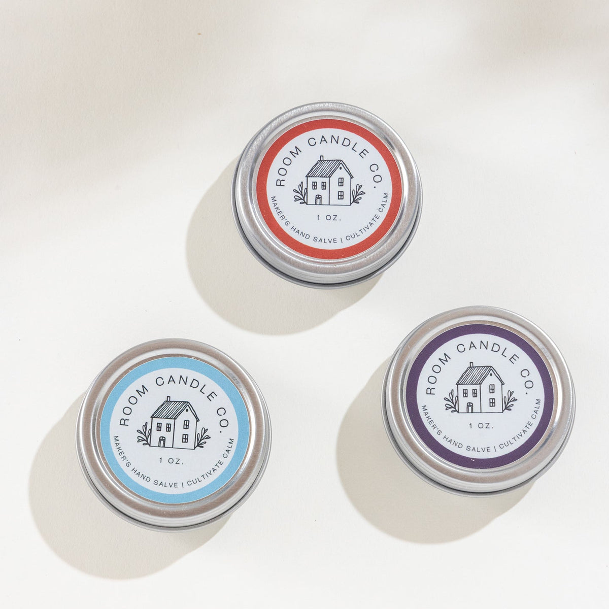Three 1 oz metal tins of Room Candle Co. Maker's Hand Salve in Lemongrass and Tea Tree, Blood Orange and Myrrh, and Black Currant and Absinthe, displayed against a plain background.
