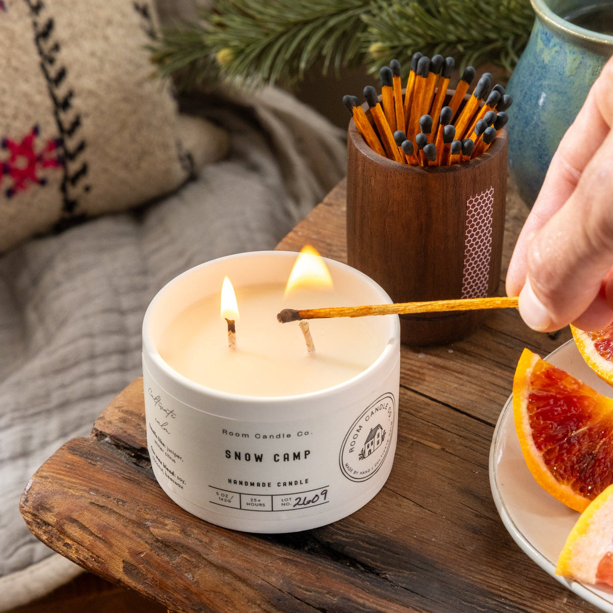 A hand lighting the Snow Camp candle by Room Candle Co. with a match, featuring blood orange and Fraser fir.